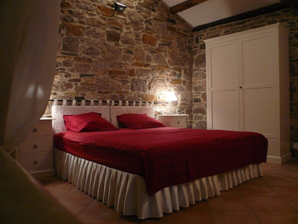 Une chambre avec un mur en pierre, un lit avec une literie rouge, une table de chevet avec une lampe et une armoire.