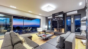 A modern living room with a large window overlooking a cityscape at dusk. There is a fireplace, a sofa set, and a coffee table.