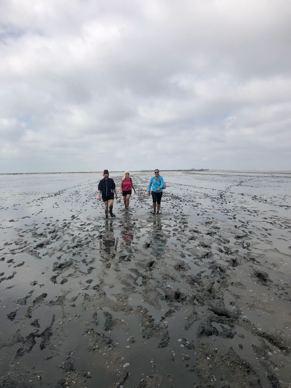 Tre personer går på en mudret strand under en overskyet himmel.