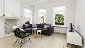 Una sala moderna con un sofá gris oscuro, una silla negra, una mesita redonda pequeña y un escritorio blanco con un monitor de computadora.