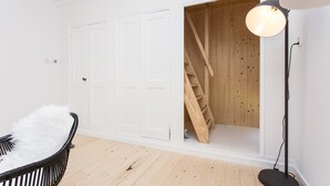 Una habitación con una puerta blanca, una escalera de madera, una lámpara de pie negra y un sillón blanco y mullido.
