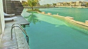A pool with a wooden deck and lounge chairs, surrounded by a calm water body and buildings in the background. - INCREDIBLE WATER FRONT APARTMENT WITH AMAZING VIEW (CAP CANA)