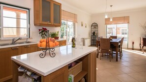 Een keuken met houten kasten, een centraal eiland met fruit en wijn, een eetgedeelte met stoelen, en een uitzicht door een raam.