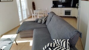 A modern living room with a grey sectional sofa, a dining table with chairs, a kitchen area with white cabinets and appliances, and a large window with curtains. - Traditional seafront ground floor apartment. Children free in summer. (Punta Mujeres)