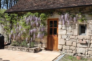 Ein Steinhaus mit Holztür und Fenster, umgeben von einem Garten mit violetten Blumen.