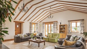 Ein geräumiges Wohnzimmer mit hoher Decke, Holzbalken und großen Fenstern. Es gibt mehrere Sitzbereiche, darunter eine Sofagarnitur, ein grüner Sessel und ein Ledersessel. Ein Holztisch steht in der Mitte, und eine Bücherwand ist an der Wand angebracht.