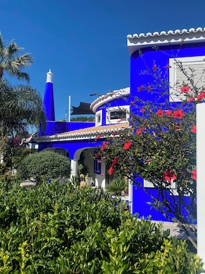 Ein blaues Gebäude mit einem weißen Turm, umgeben von Grünpflanzen und roten Blüten.