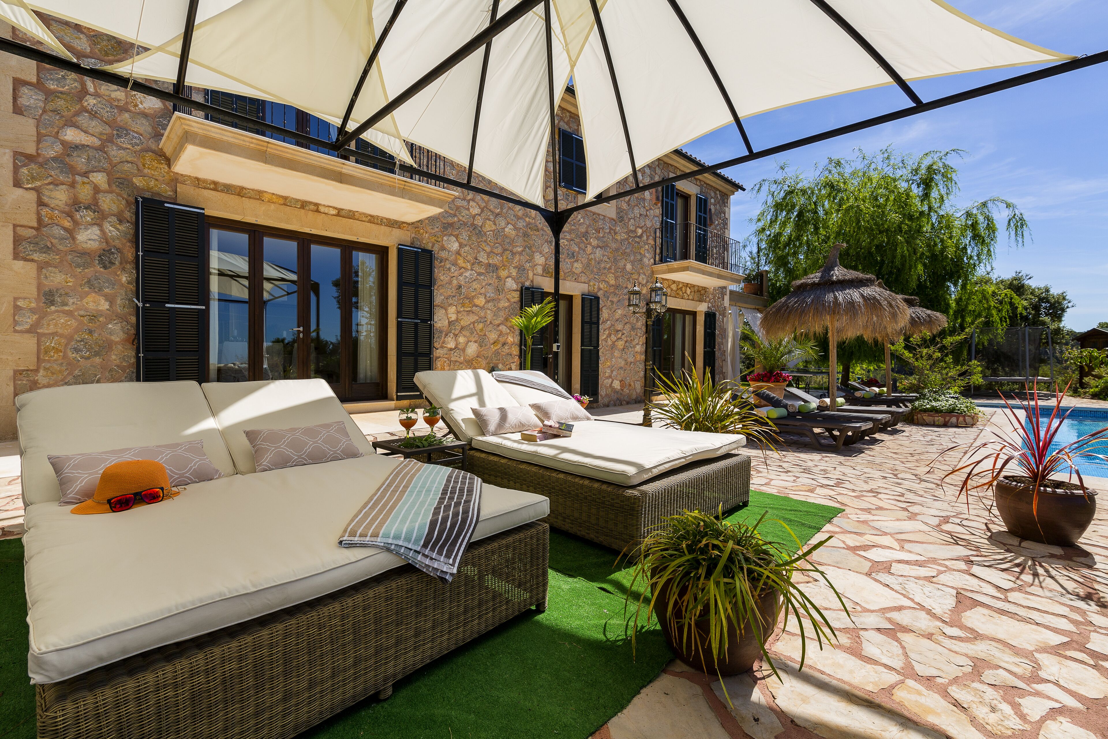 A sunlit outdoor lounge area with wicker furniture, a pool, and a stone building facade.