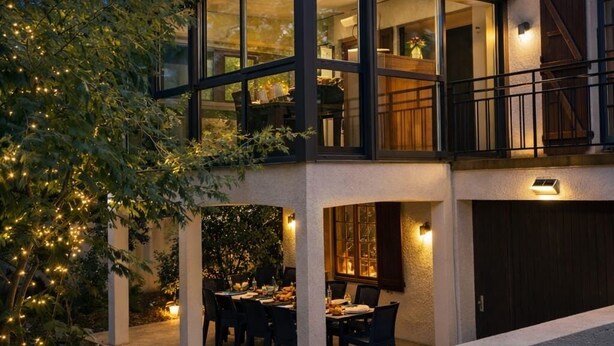 Ein modernes, zweistöckiges Haus mit einem gläsernen Obergeschoss, einem Essbereich inklusive Tisch und Stühlen sowie einer überdachten Außenlounge.