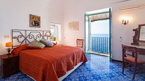 A bedroom with a bed, a chair, a desk, a nightstand, a lamp, a picture frame, a chandelier, and a view of the sea.