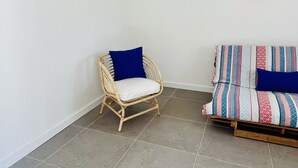 A room with a wicker chair, a sofa with a colorful blanket, and three beach-themed paintings on the wall. - Salty Soul Beach House (Costa da Caparica)