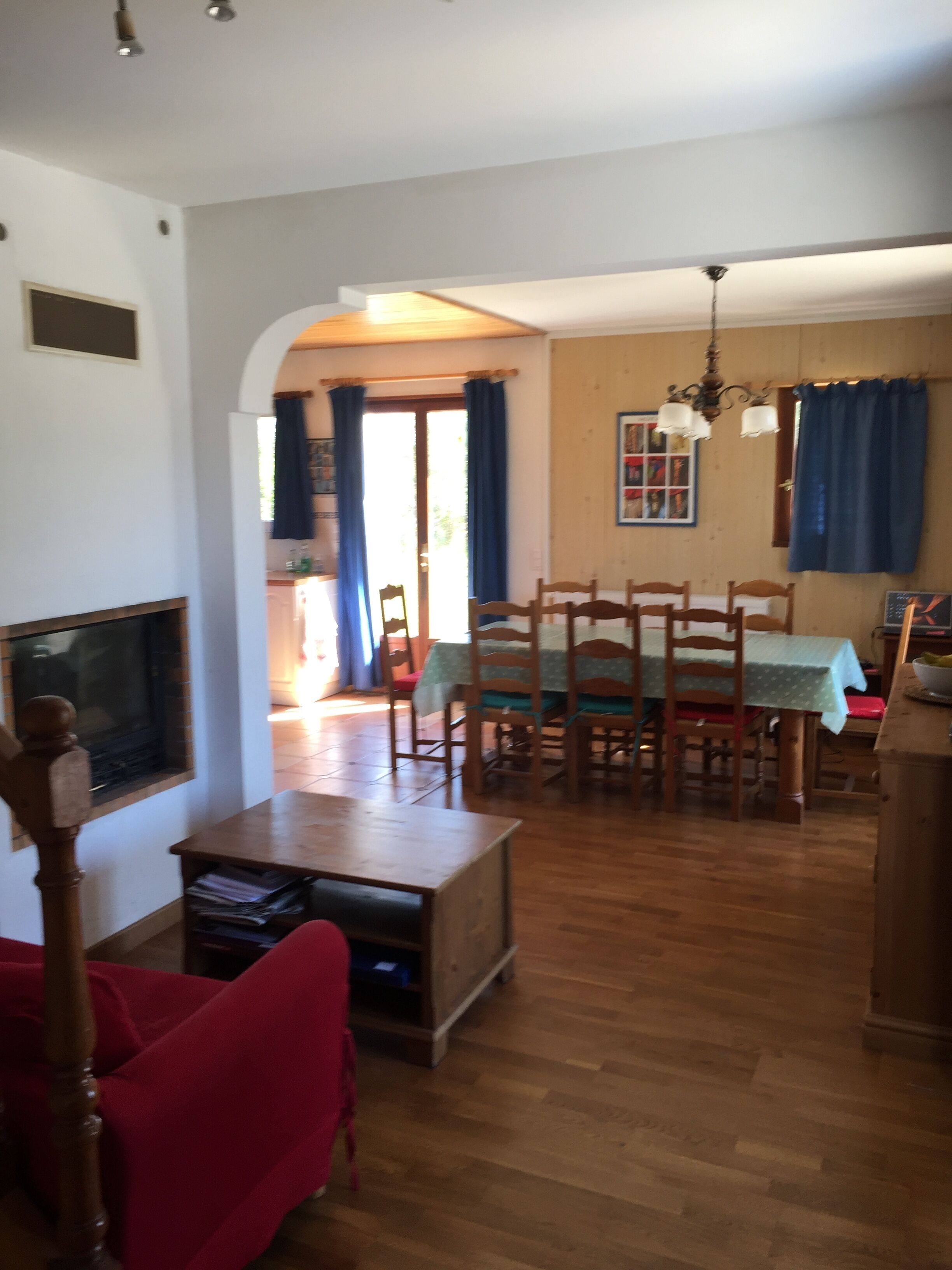 A living room with a wooden coffee table, a red armchair, and a TV mounted on the wall. There is a dining area with a table and chairs, and a kitchen area with a sink and countertop visible.