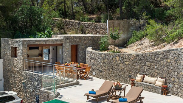 Ein Gebäude mit Steinwänden, das eine Küche, einen Essbereich und eine Poollounge mit Liegestühlen und einem kleinen Tisch bietet.