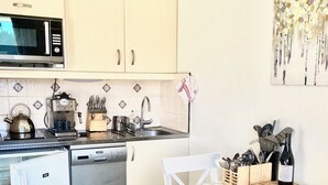 A compact kitchen with a microwave, kettle, coffee maker, and dishwasher. There is a dining table with a patterned tablecloth and white chairs. A wine bottle is on the table, and a towel hangs on the wall. - Ski Apartment With Spectacular Mountain View (Flaine Forêt) @flaine1969 (Arâches-La-Frasse)