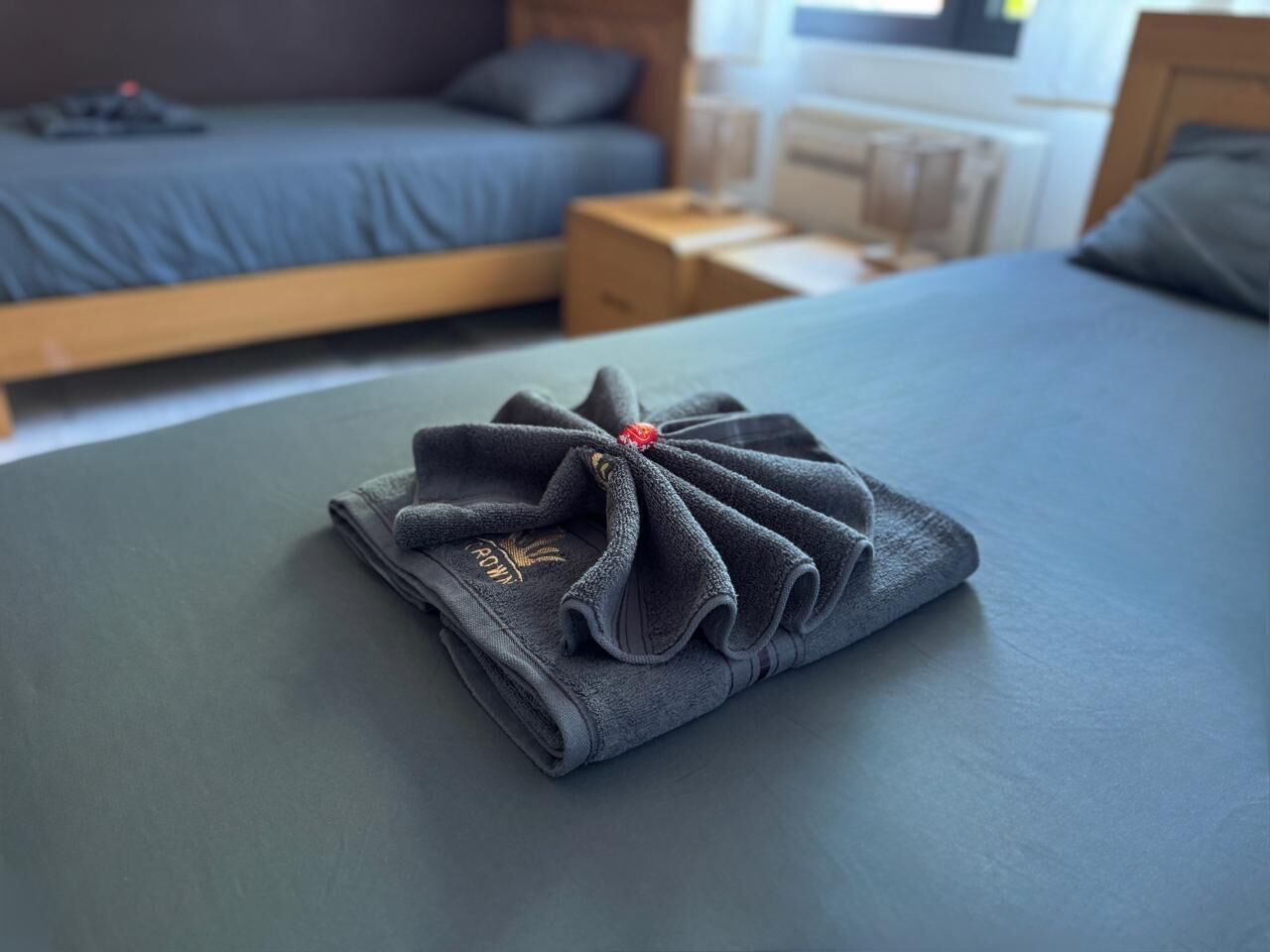 A neatly made bed with a folded towel on top, a wooden headboard, and a nightstand with a lamp.