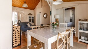 Comedor con techo de madera, una mesa blanca y sillas. Se ve una cocina a través de una puerta abierta.