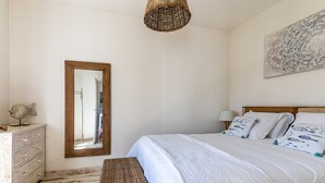 Un dormitorio con una cama grande, un aparador de madera, un banco de mimbre, un espejo y una lámpara colgante.
