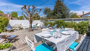 Una terraza de madera con una mesa lista para comer, rodeada de sillas de lounge y una cerca blanca de piquetes.