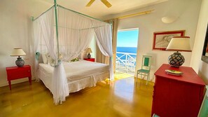 A bedroom with a four-poster bed, a red nightstand, a red dresser, and a view of the sea.
