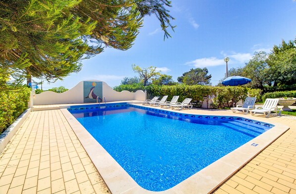 A rectangular swimming pool with clear blue water, surrounded by a tiled deck. There are lounge chairs and a blue umbrella on the deck.