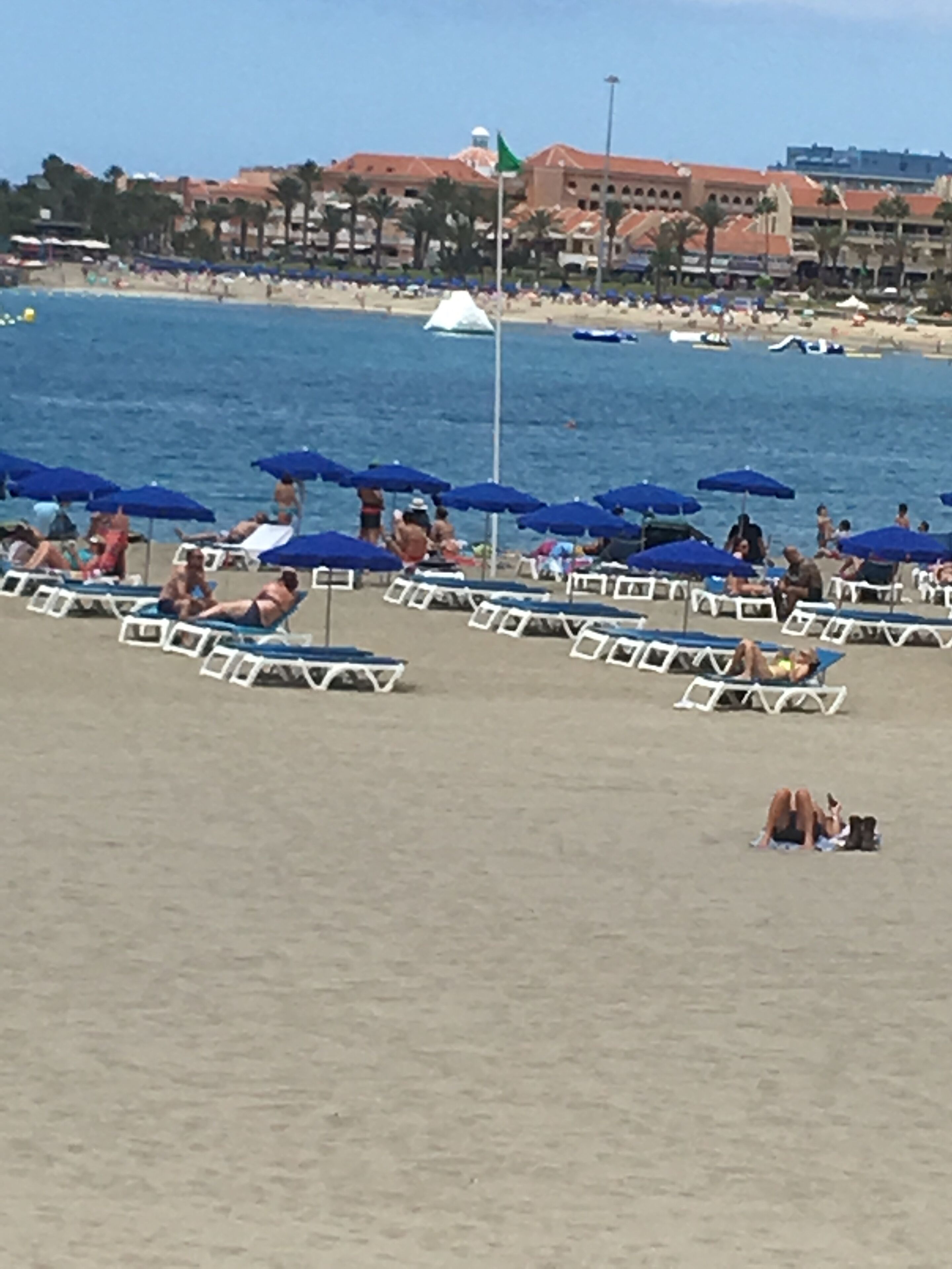 A beach with rows of sun loungers and umbrellas, a flagpole with a flag, and buildings in the background.