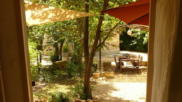 Une porte ouverte donne sur un patio doté d’une terrasse en bois, d’une table et de chaises, le tout entouré de verdure.