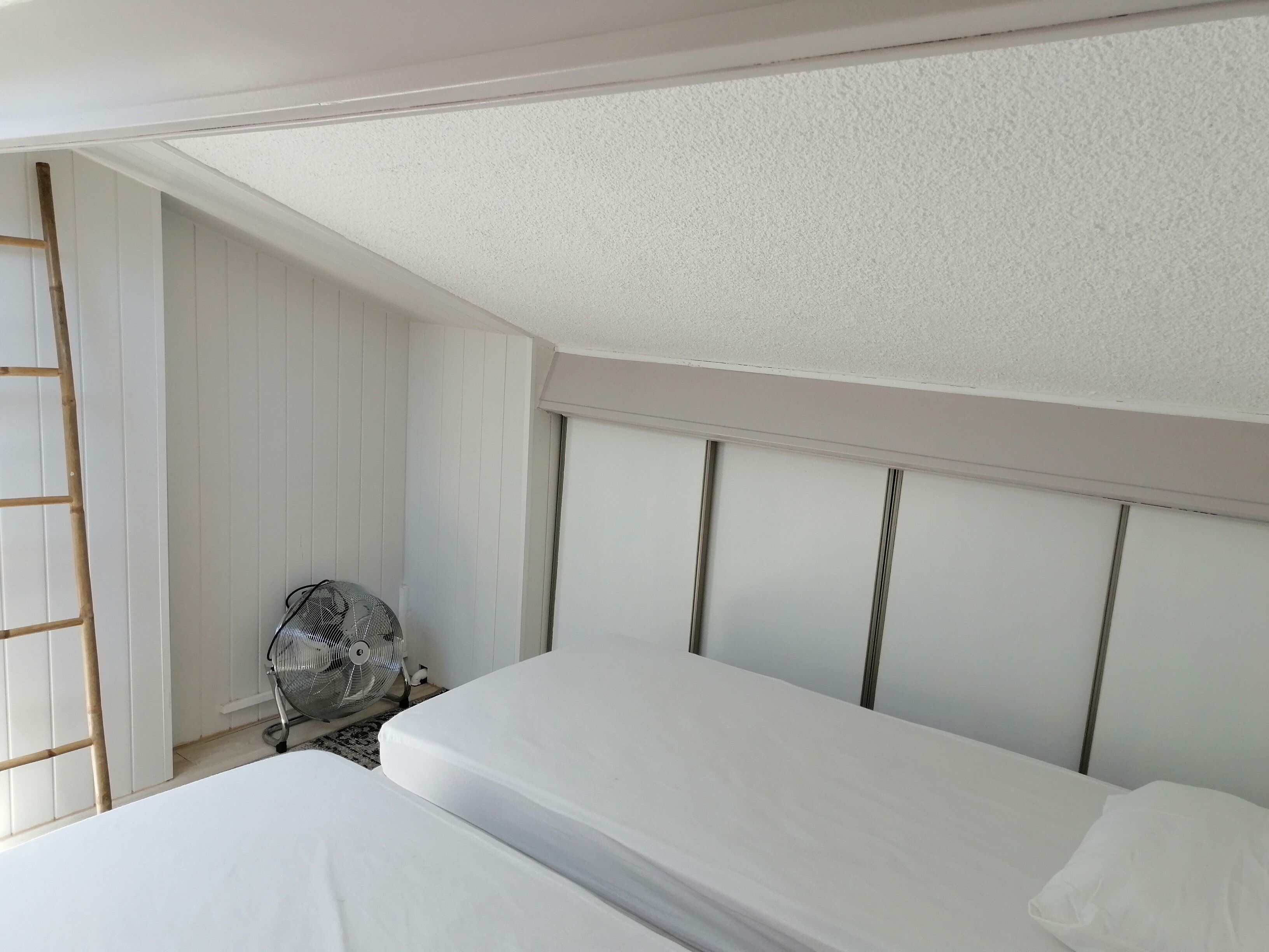 Quarto com duas camas, paredes brancas, escada de madeira e um ventilador.