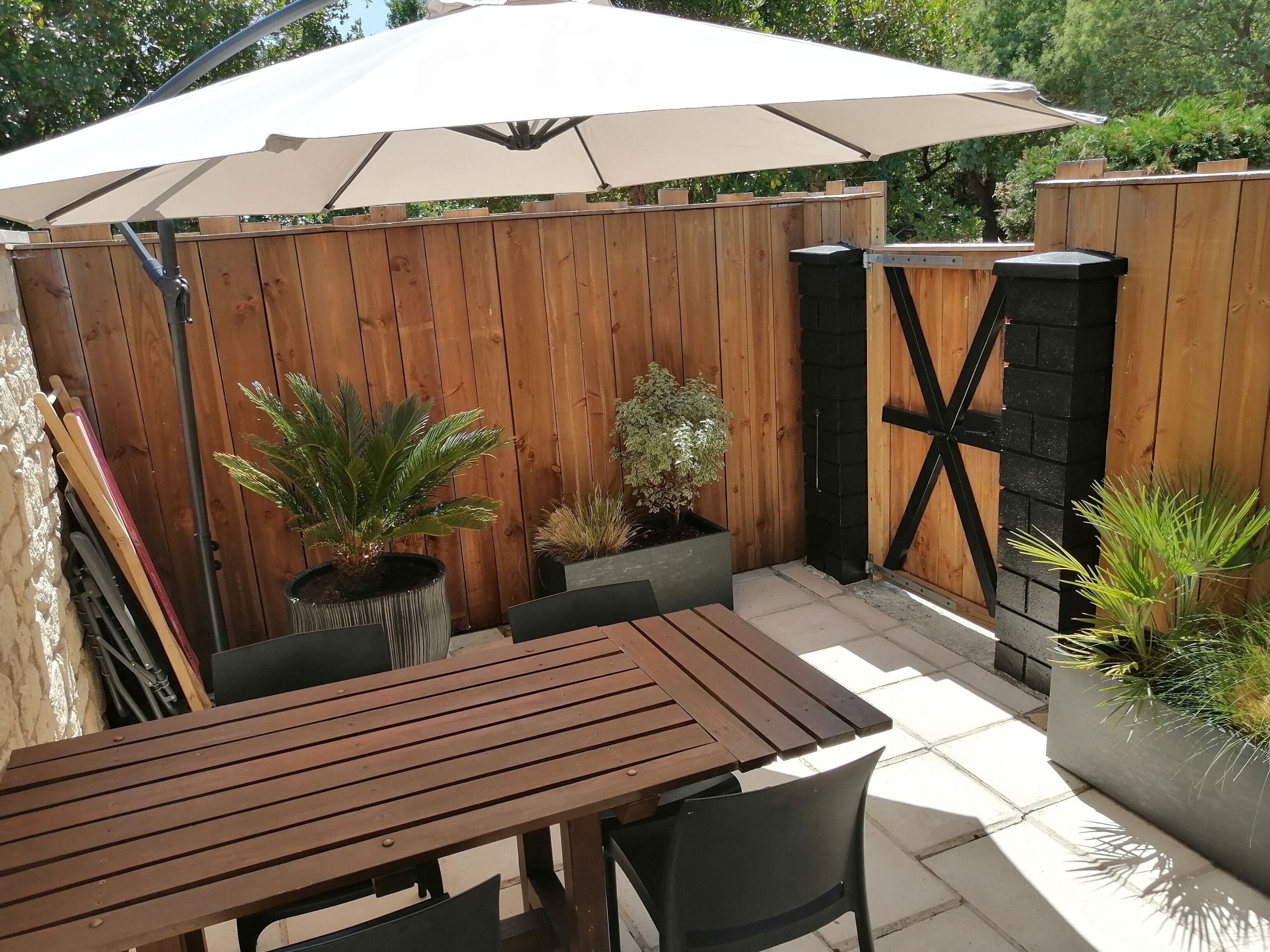 Een houten terras met een tafel en stoelen, omgeven door potplanten en een grote parasol.