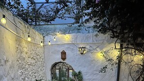Un patio exterior acogedor con una pared de piedra rústica, plantas en macetas, una silla de mimbre e iluminación con luces colgantes.