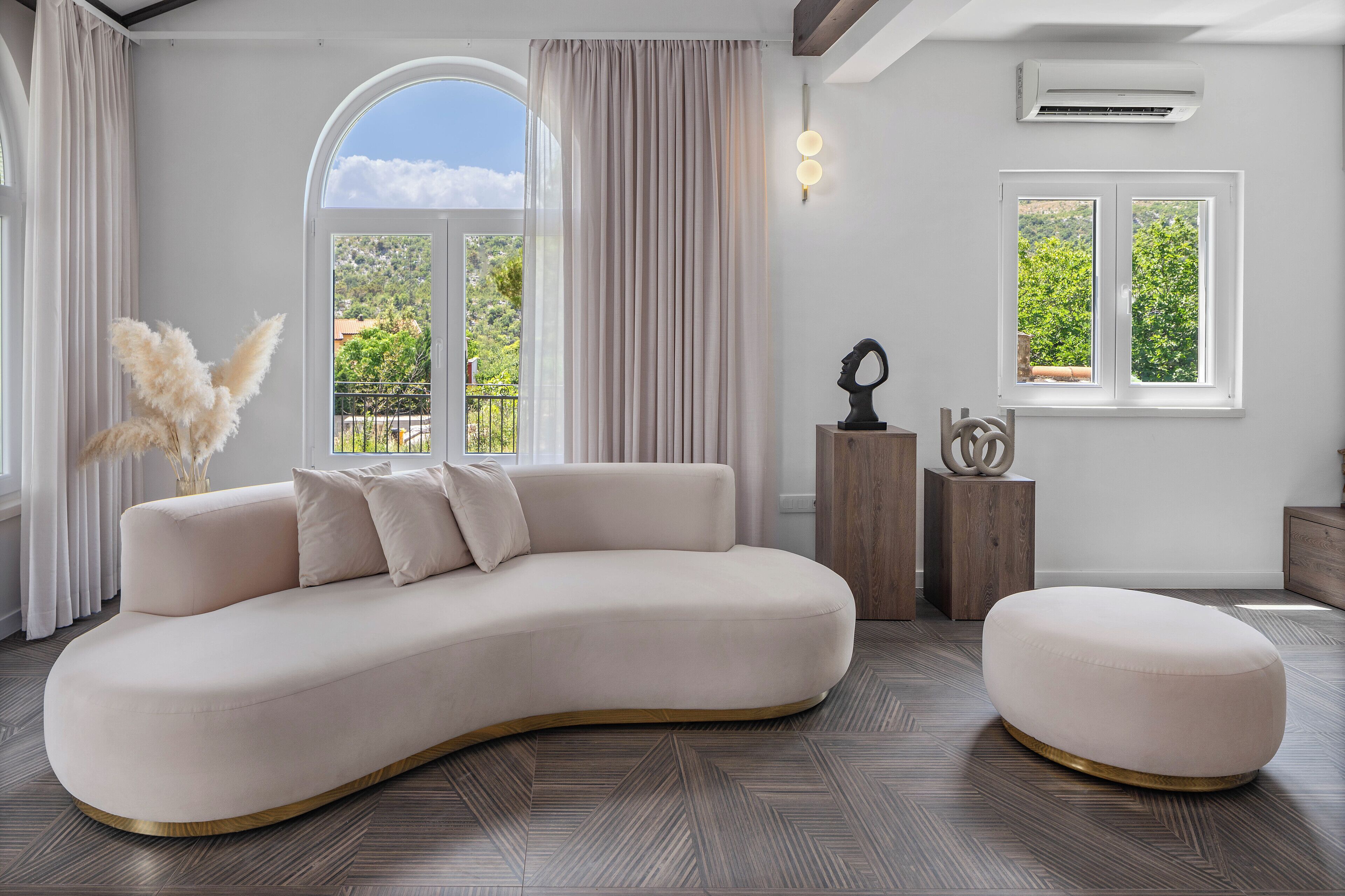 A modern living room with a curved sofa, an ottoman, a decorative vase, and a view of greenery through the windows.