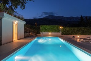 A rectangular swimming pool with a clear blue water surface, surrounded by a stone wall and a metal railing. There are steps leading up to a door on the left side, and a green hedge in the background.