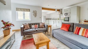 A modern living room with a leather sofa, a wooden coffee table, and a grey couch with colorful pillows. A kitchen with white cabinets and stainless steel appliances is visible in the background. - Luxury Cottage | New Forest | Family | Friends | (Redlynch)