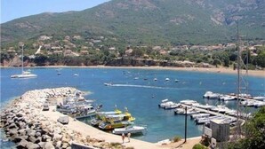 Una marina costera con barcos amarrados, un rompeolas rocoso y montañas al fondo.