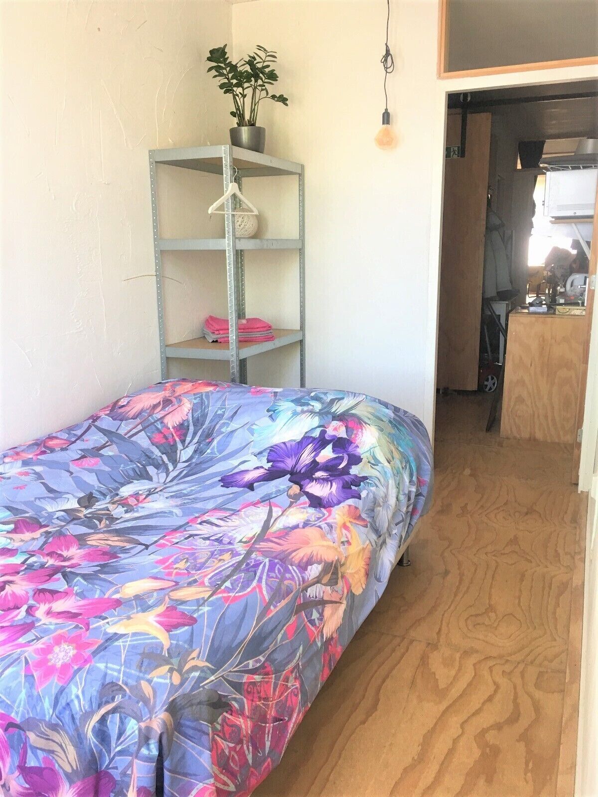 Un dormitorio con una colcha estampada, una repisa metálica con ropa doblada y una planta en maceta.