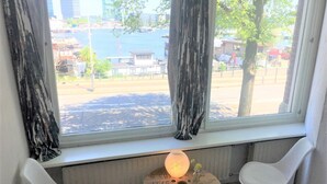 A room with a large window overlooking a body of water, two white chairs, a small round table with a lamp, and a floral-patterned bedspread.