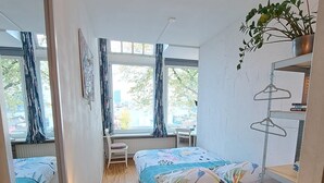 A compact bedroom with a bed, a small desk, and a chair. There are windows with curtains, a plant on a shelf, and a mirror on the wall.