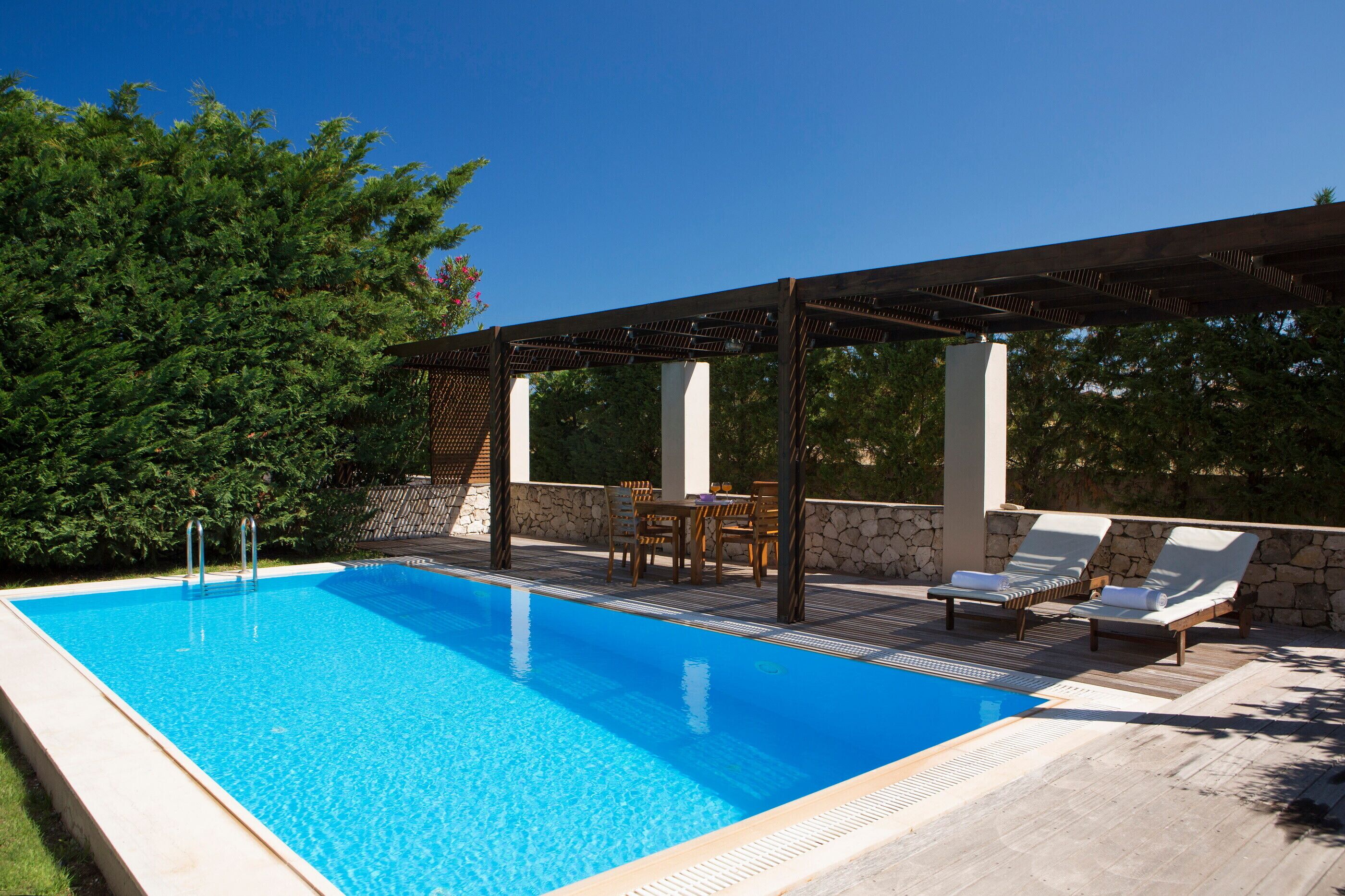 Una piscina rectangular con agua azul cristalina, rodeada por un suelo de madera y muros de piedra. Hay sillones y una mesa bajo un pérgola.