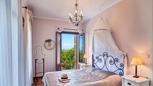 A bedroom with a bed, a nightstand, a chandelier, a window with a view, and a curtain. - Aeriko-Varko Beachfront Villa with Stunning View and Private Pool (LEFKADA)