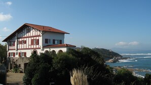 Un bâtiment à deux étages, doté d’une façade blanche et d’un toit recouvert de tuiles rouges, qui surplombe une zone côtière et offre une vue sur la mer et un paysage vallonné.