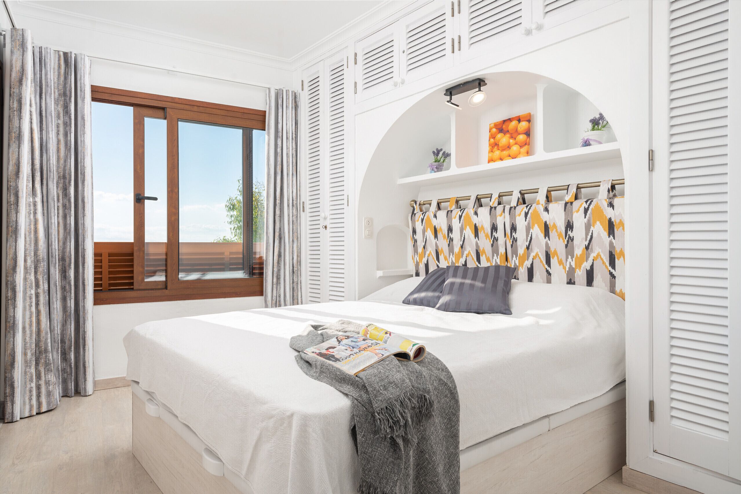 Een moderne slaapkamer met een groot bed, een raam met gordijnen, een inbouwkast met decoratieve objecten en een plafondventilator.