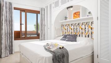 Een moderne slaapkamer met een groot bed, een raam met gordijnen, een inbouwkast met decoratieve objecten en een plafondventilator.