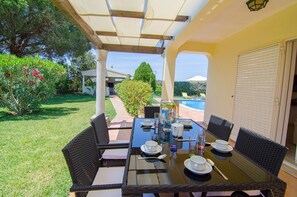 Um pátio externo com mesa posta para refeições, cercado por cadeiras de vime, e uma área coberta com vista para uma piscina e vegetação.