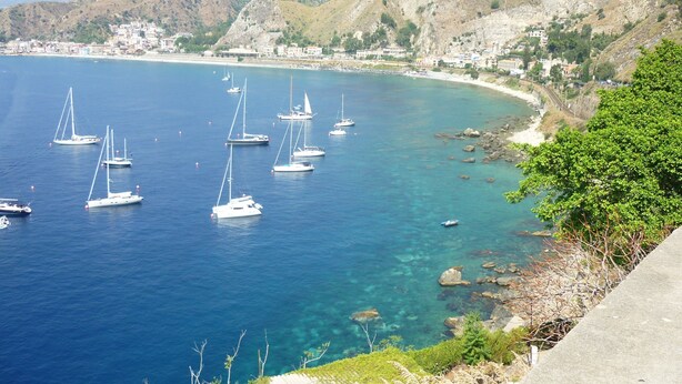A coastal area with clear blue waters, several sailboats, and a rocky shoreline with greenery.