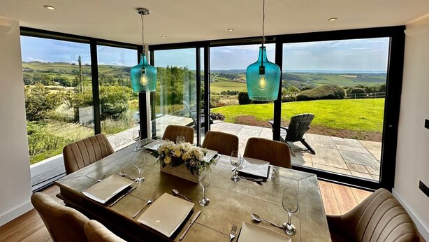 Comedor con una mesa dispuesta para seis personas, ventanas amplias que ofrecen vistas al campo y dos lámparas colgantes.
