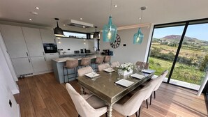 Una cocina moderna con una isla central, un comedor con mesa y sillas de madera, y ventanas grandes que ofrecen vistas al campo.