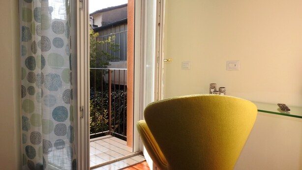 A room with a yellow chair, a glass shelf with a remote control, and a view of a balcony with plants and a building in the background. - Completely renovated apartment in the center (Ravenna)