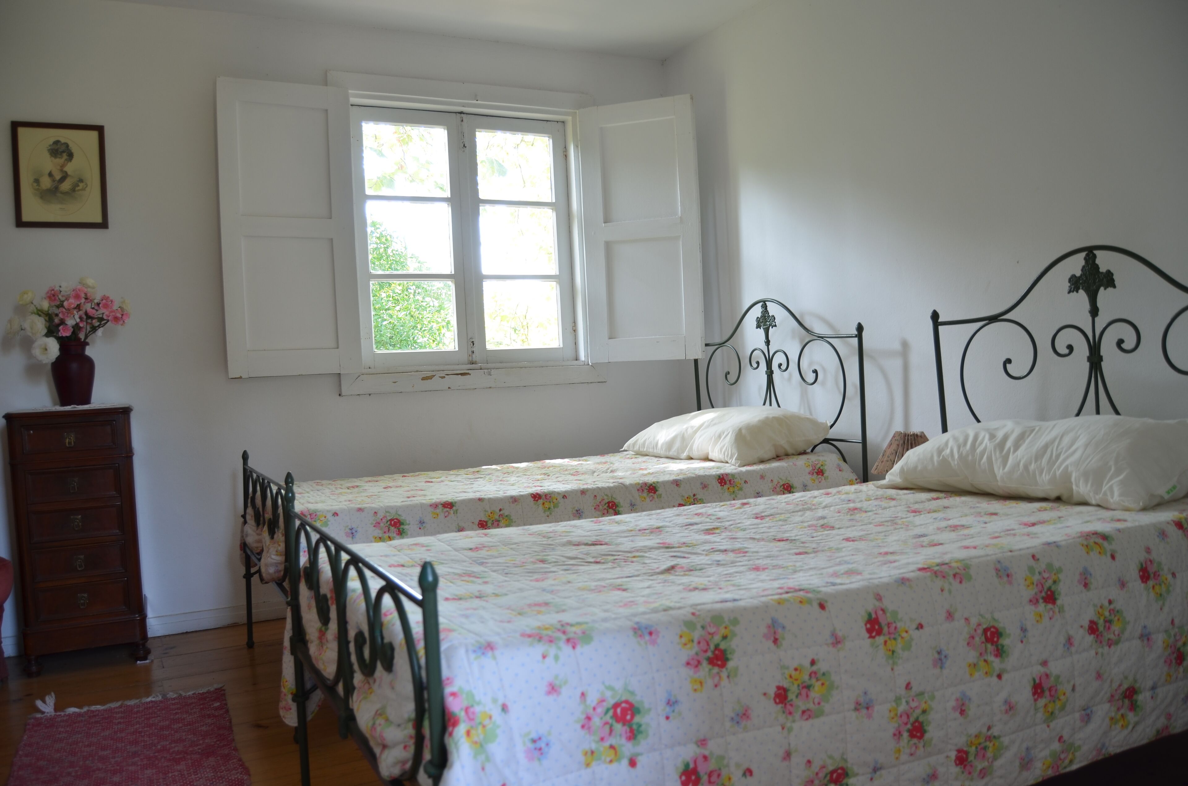 Une chambre avec deux lits simples, une commode, une fenêtre avec des volets blancs et un tableau encadré au mur.