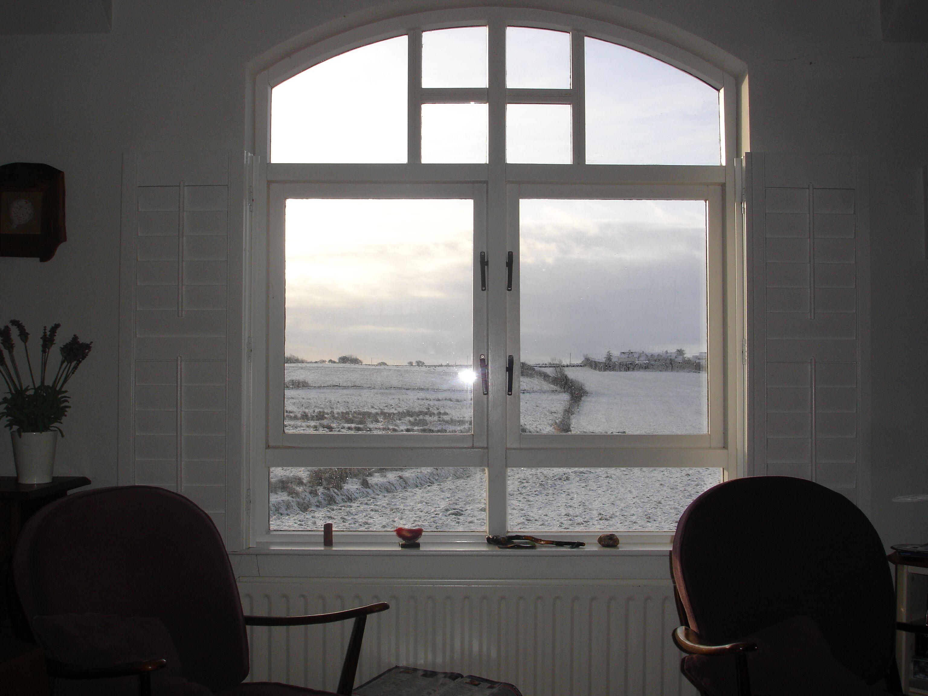 A room with a large arched window overlooking a snowy landscape, two chairs, and a radiator.