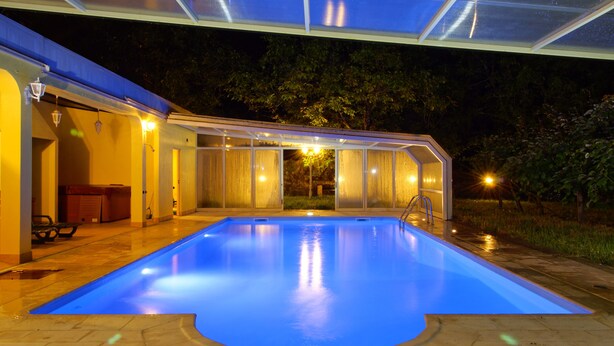 Une piscine dotée d’une structure en verre, entourée d’un patio aménagé avec des sièges d’extérieur et une entrée couverte.