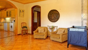 Un salon chaleureux avec un canapé, des fauteuils et une table basse. On aperçoit une cuisine à l’arrière-plan, avec des meubles en bois et un lave-linge.